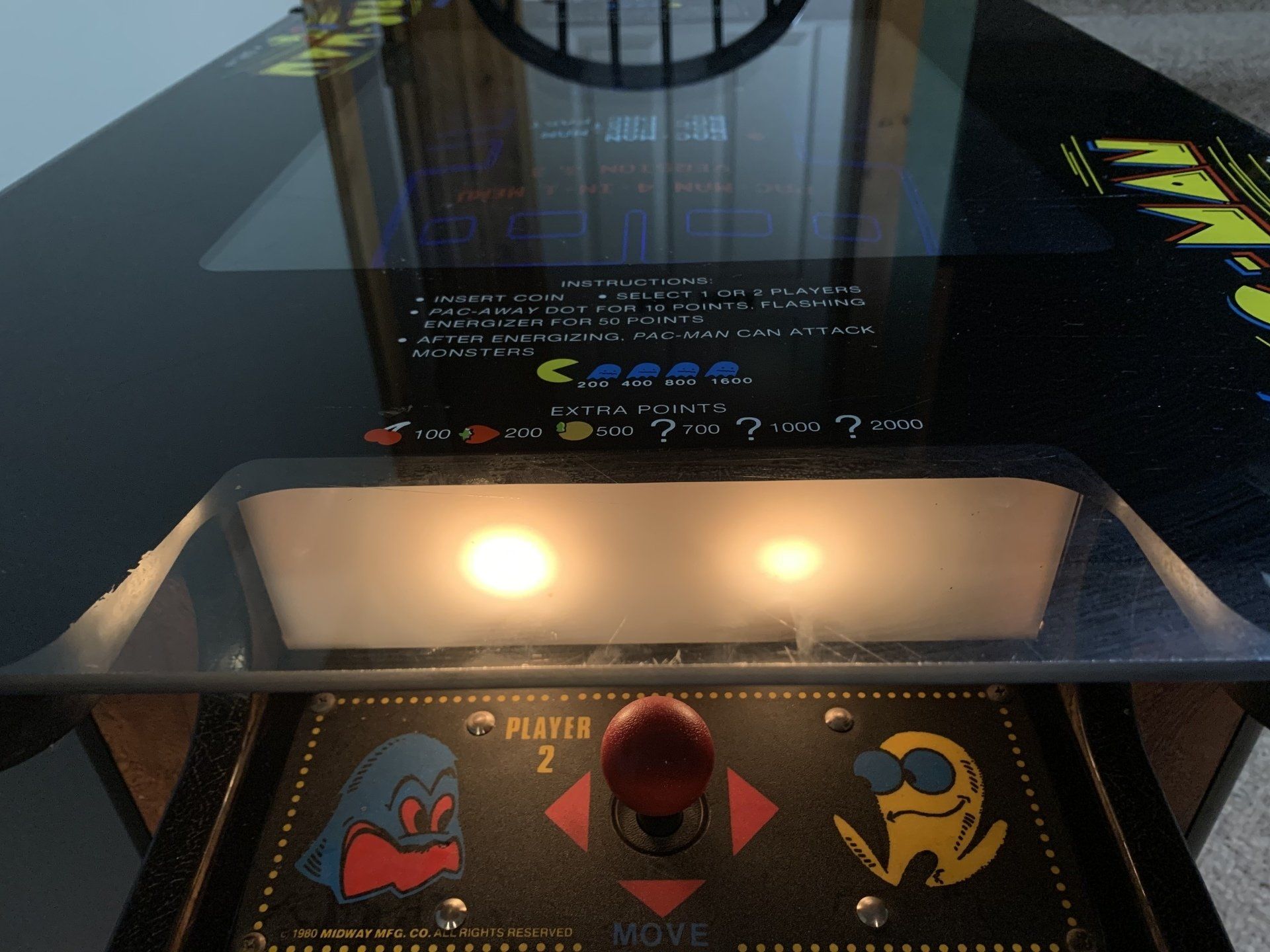 Arcade game, close-up. Pac-Man control panel with joystick and buttons; illuminated marquee with game title.