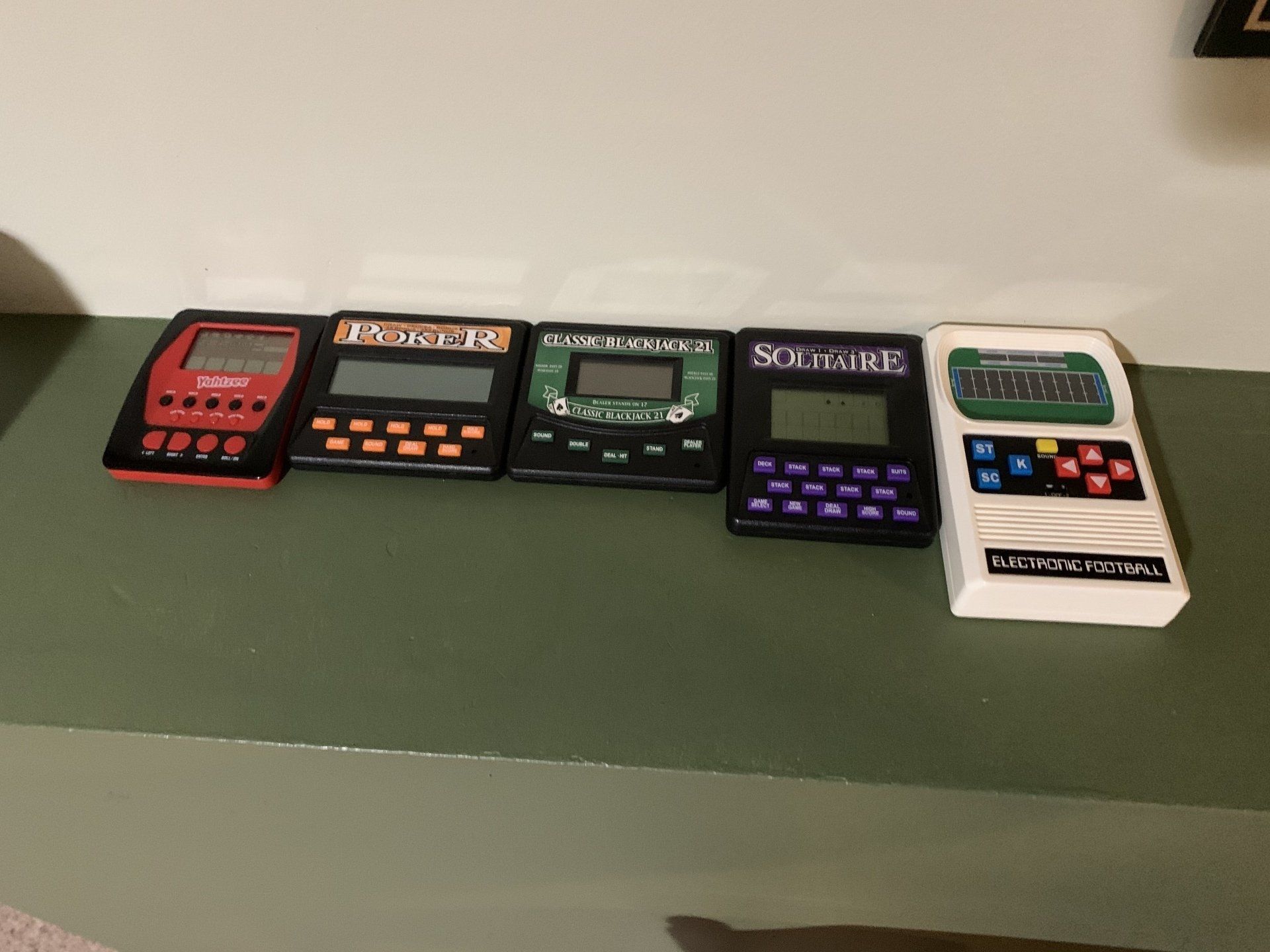Five handheld electronic word games of varying colors on a green surface.