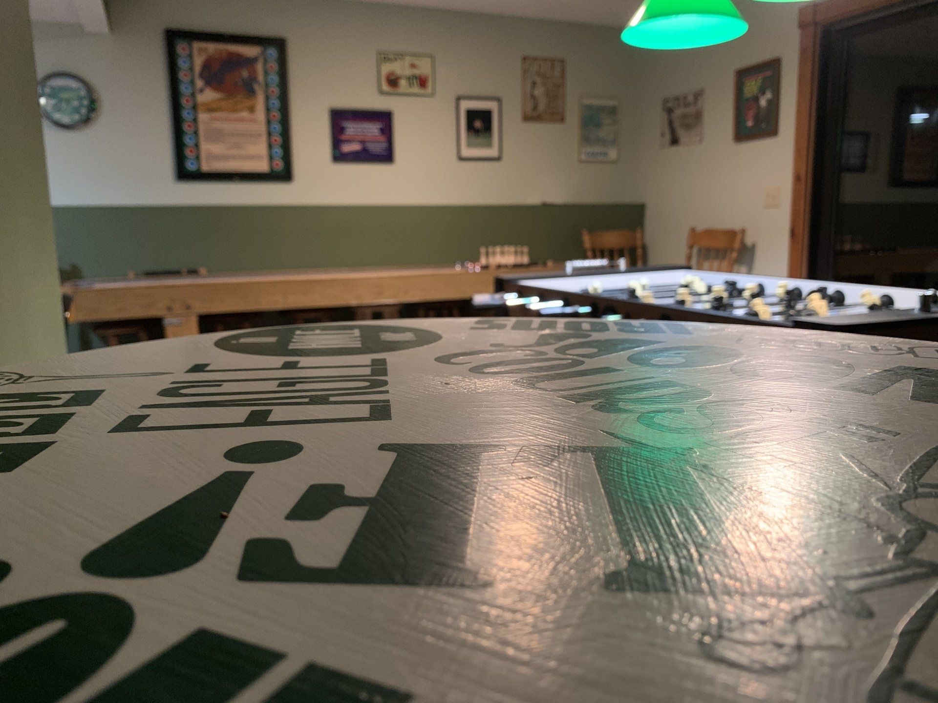 A room with shuffleboard and other game tables, posters on the wall, and a green-lit ceiling fixture.