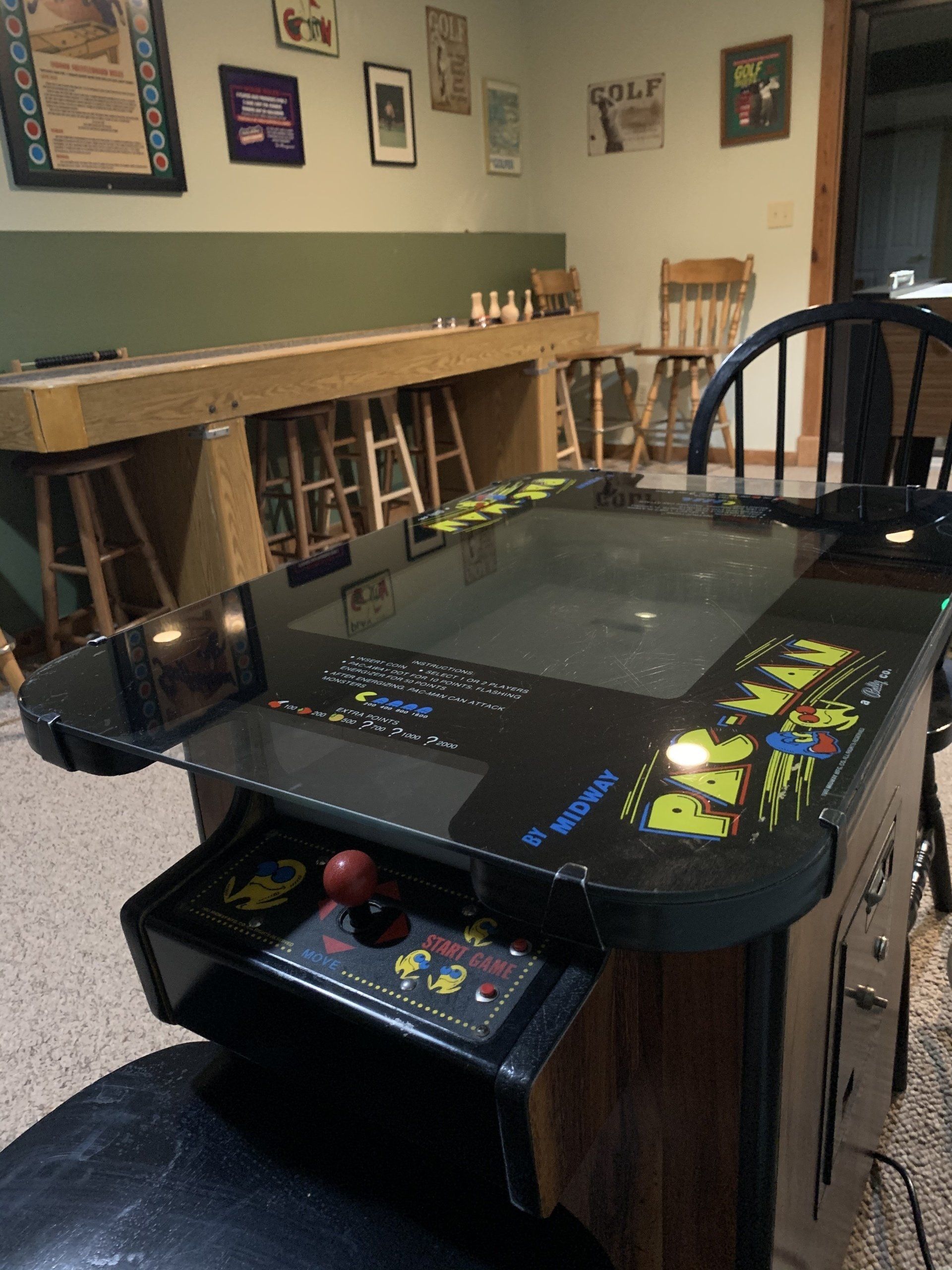 Pac-Man arcade game in a game room with a shuffleboard table and bar stools.
