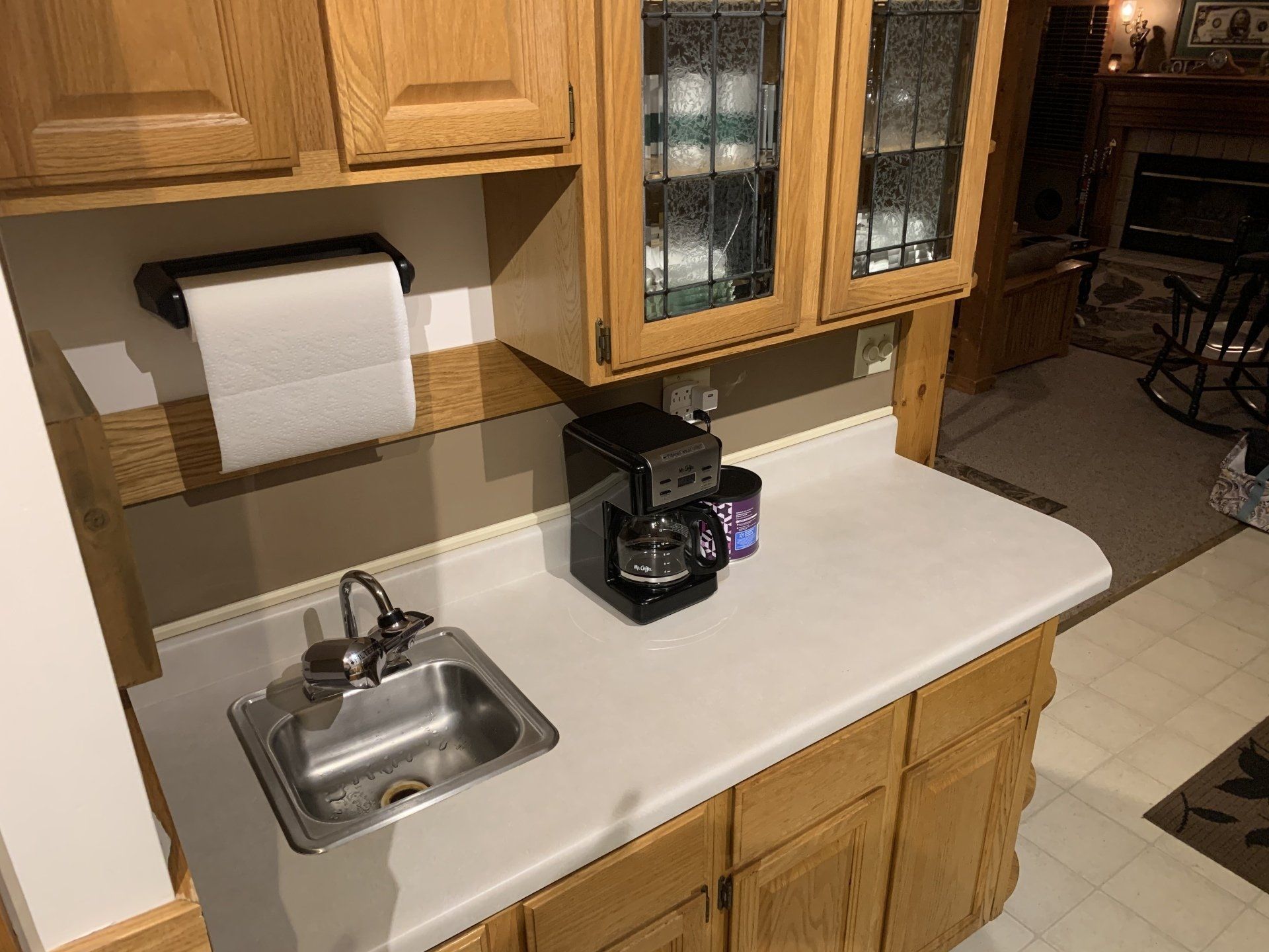 Kitchen counter with a sink, coffee maker, and cabinets. A roll of paper towels is on a shelf.
