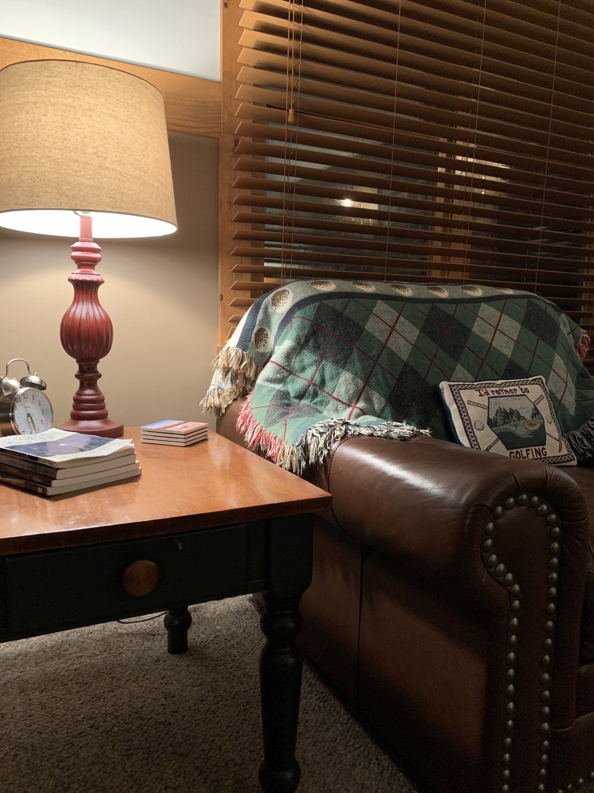 Cozy room corner: lamp on table, leather chair with blanket, wooden blinds, warm lighting.