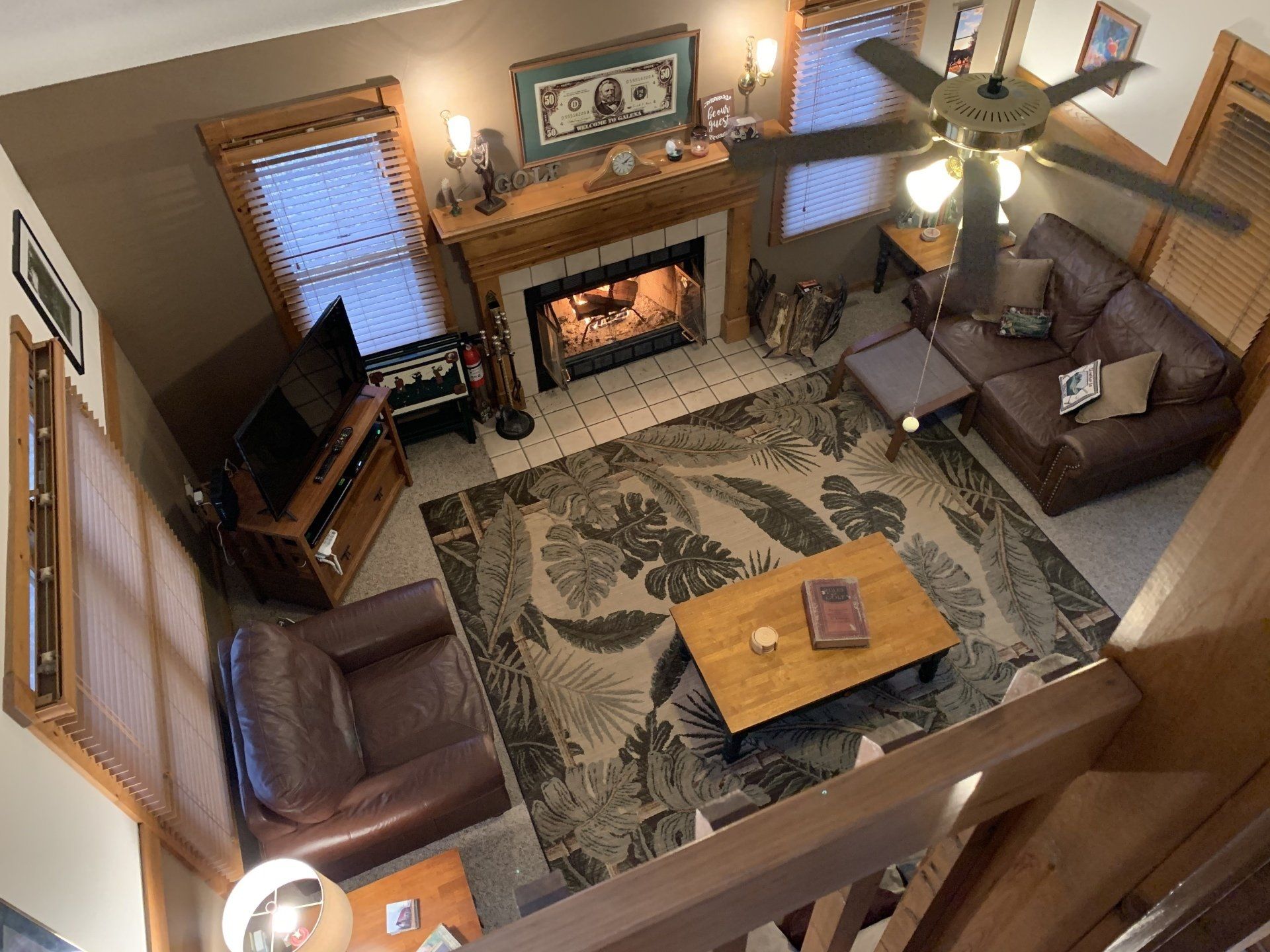 Overhead view of living room: fireplace, brown leather furniture, patterned rug, wooden coffee table, ceiling fan.