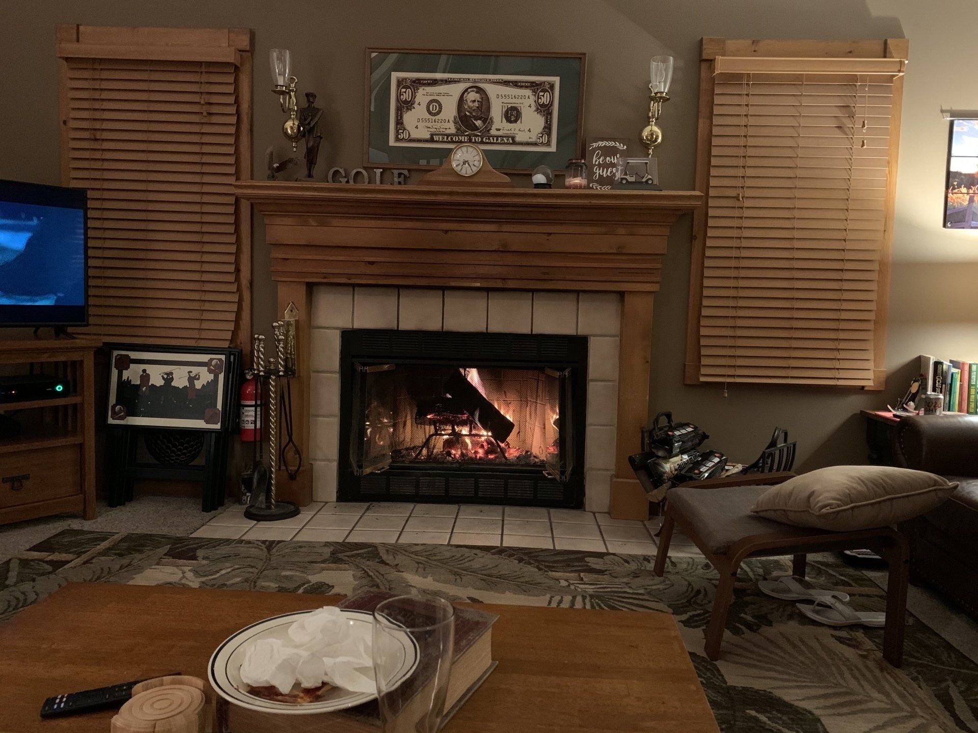 Fireplace with fire burning, mantle with decorations, blinds on either side, and a living room.