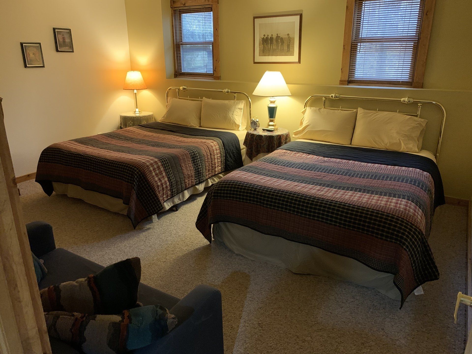 Two beds with brown comforters in a room with a small couch, lamps, and windows.