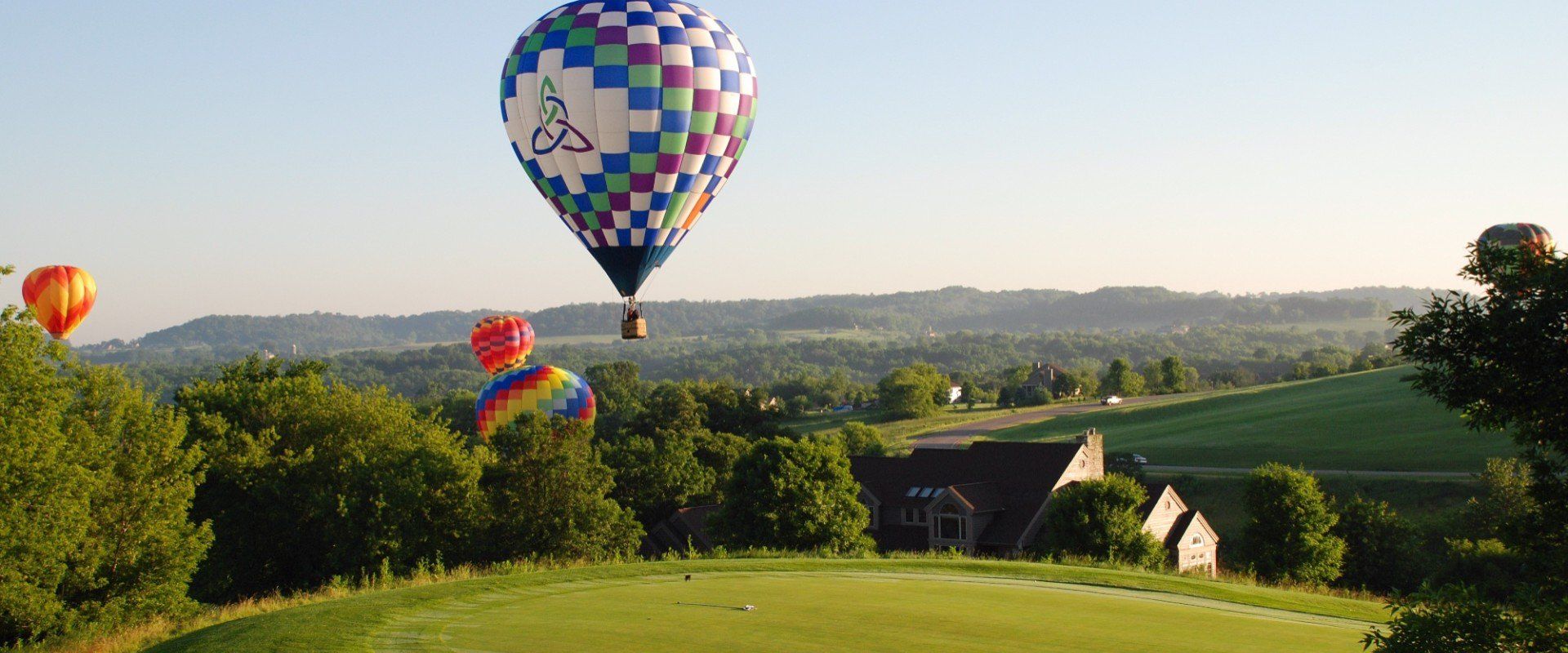 Hot air balloons over a green field with trees and a house, mountains in the distance.