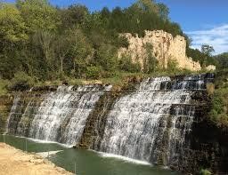 Waterfall cascading over layered rock formations, with a rocky cliff in the background and a lush forest.