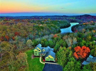 Green house with black driveway surrounded by colorful autumn trees and a lake at dusk.