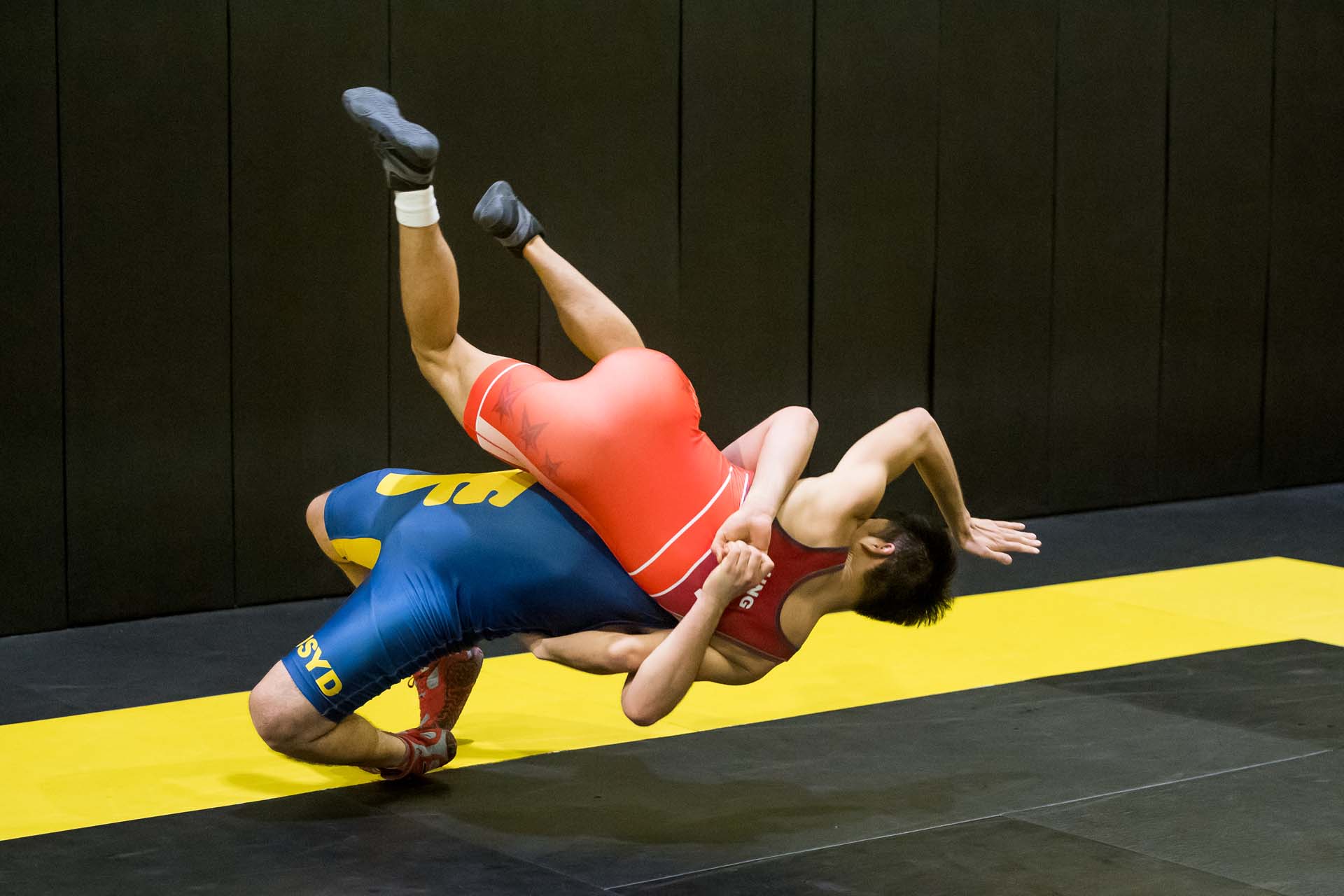 Two wrestlers grapple on a yellow mat, one in blue and one in red, in a gym.