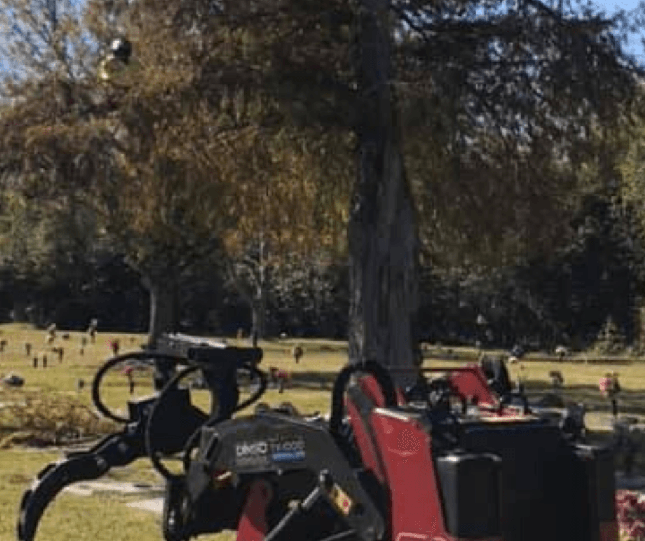 A red tractor is parked in a cemetery next to a tree.