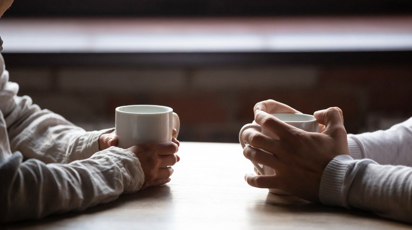 Two people sitting at a table holding white mugs, facing each other in a dimly lit setting.
