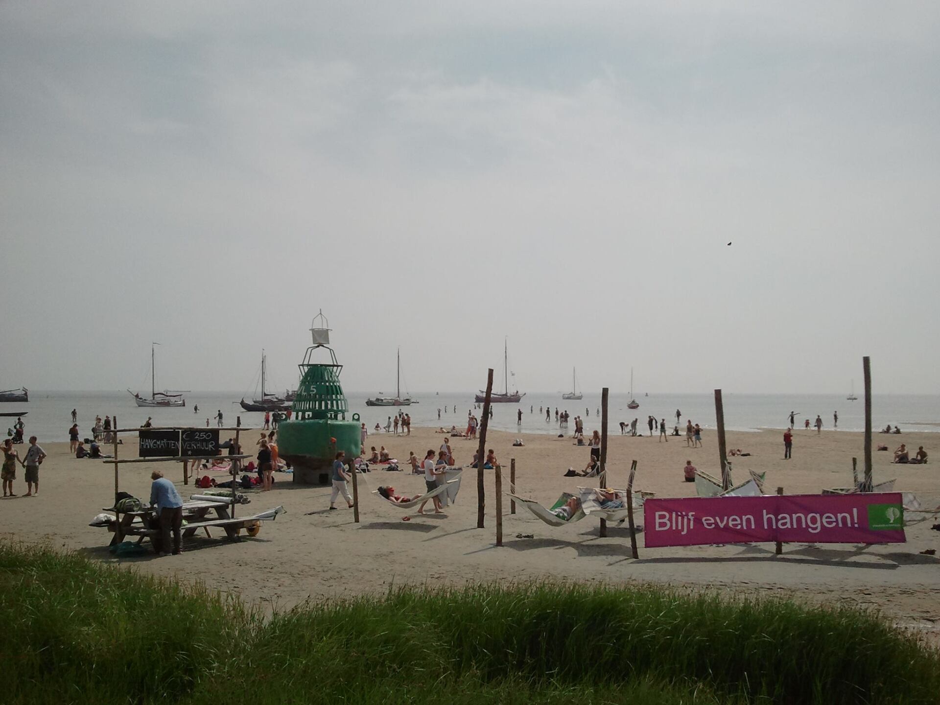 Relaxed beach scene on the Dutch Wadden Sea with people, small sailboats, a green buoy and hammocks.