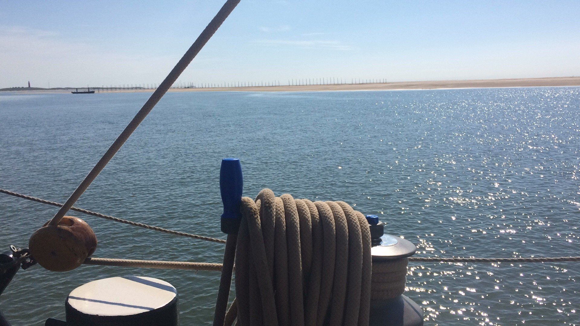 Coiled rope on deck with sparkling water and a Dutch coastline in the background under a bright sky.