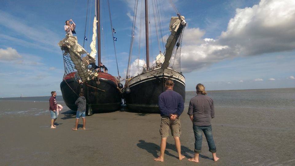 Two traditional sailing ships dried out on the Wadden Sea mudflats, with people walking around at low tide.