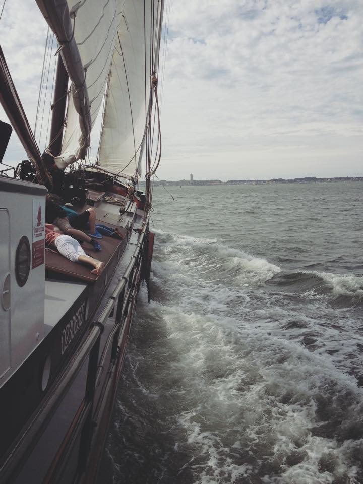 Traditional Dutch sailing ship cutting through waves, with guests on deck and a coastline on the horizon.
