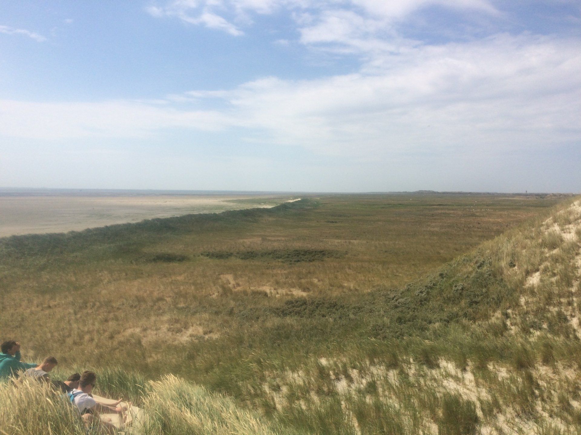 Dune slope overlooking calm water, with people sitting sheltered from the wind on a Dutch Wadden Island.