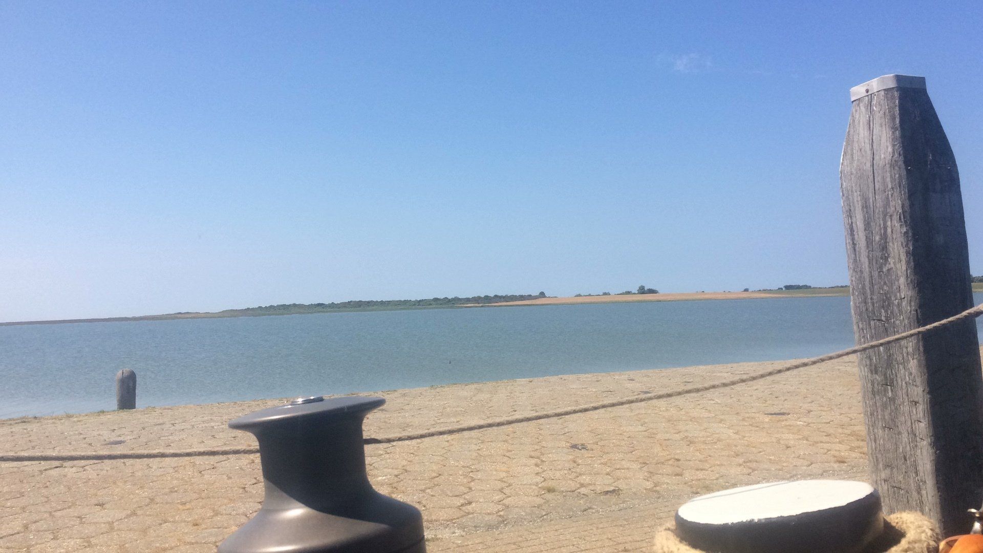 Calm beach scene with wooden posts and rope, blue sky above the water in the Dutch Wadden Sea.