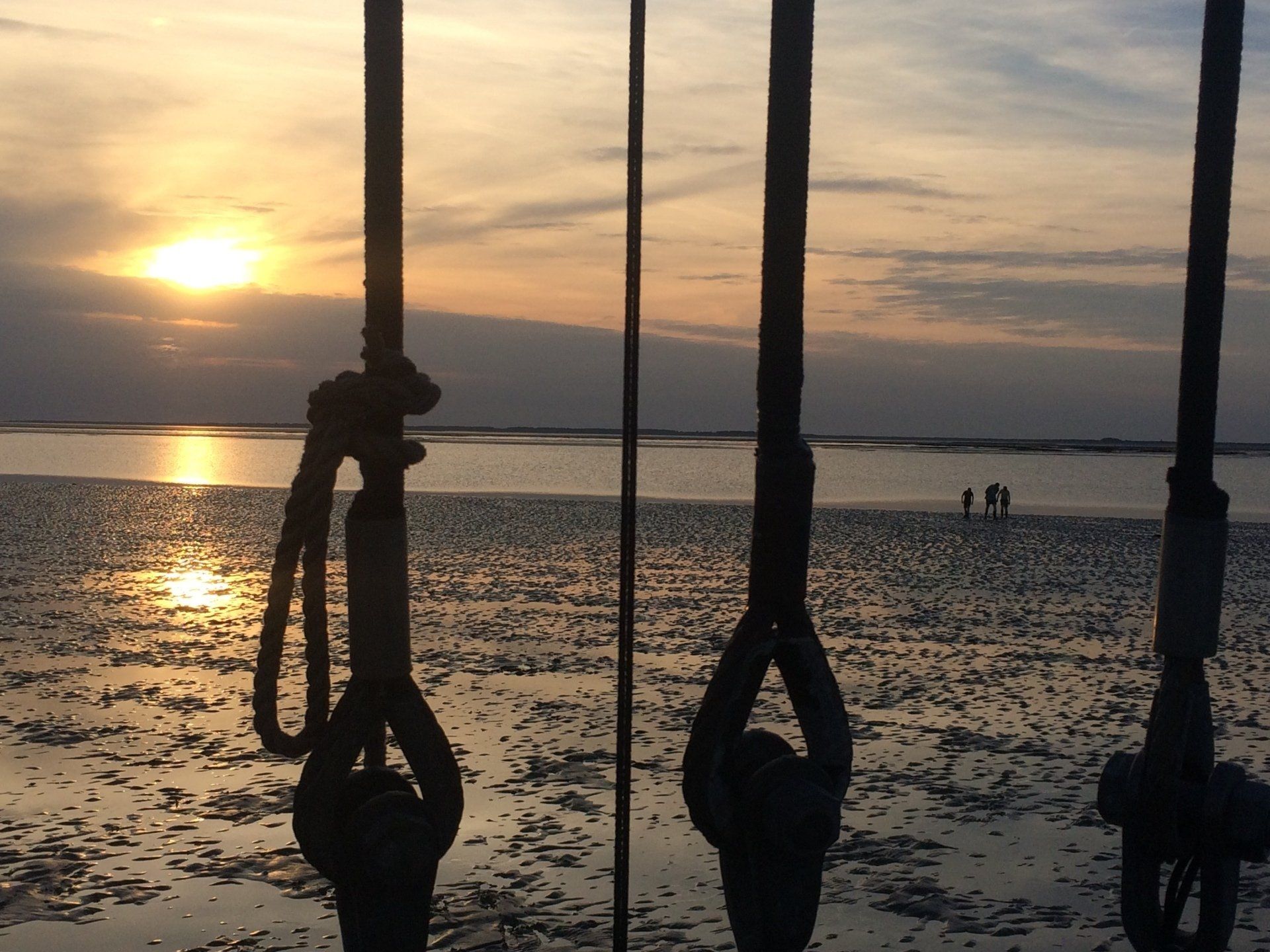 Sunset through rigging lines with people walking on wet sand and reflections on the Dutch Wadden Sea.