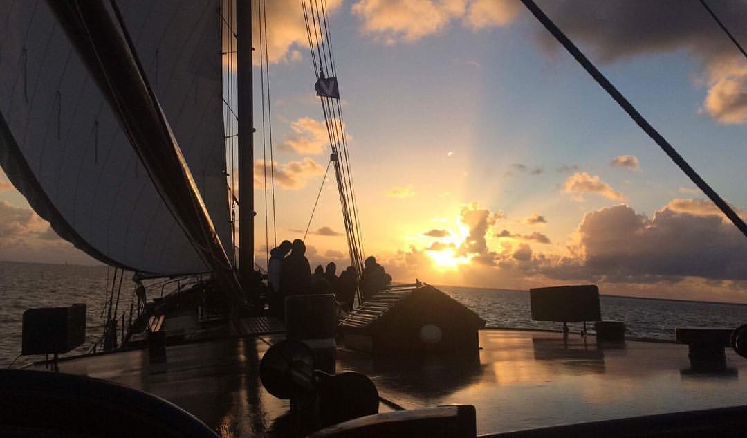 Deck of a traditional sailing ship at sunset, silhouettes against a golden sky over the Netherlands.