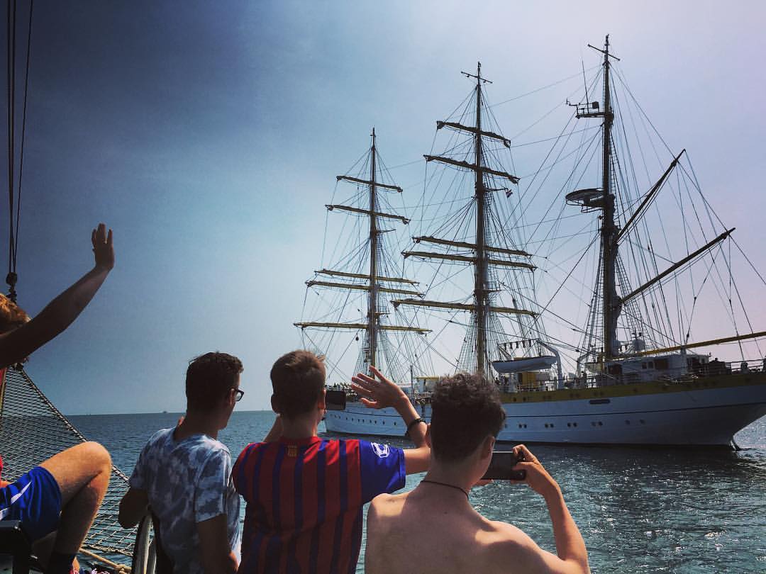 People waving from a small boat to a large traditional sailing ship on sunny Dutch waters.