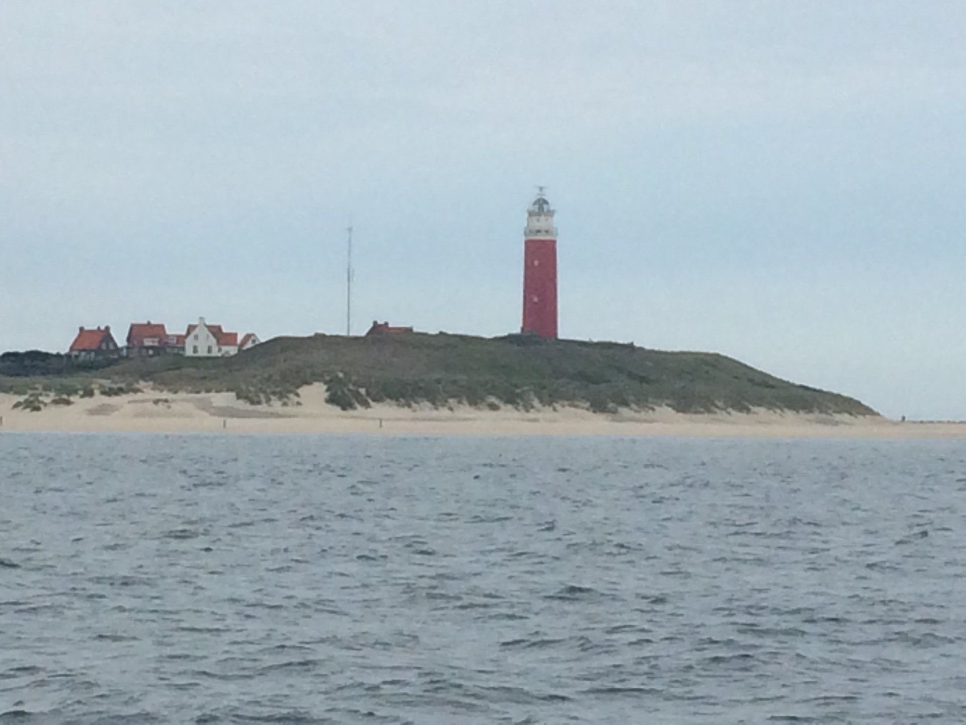 Lighthouse on a dune with buildings, seen from the sea under cloudy skies in the Netherlands.