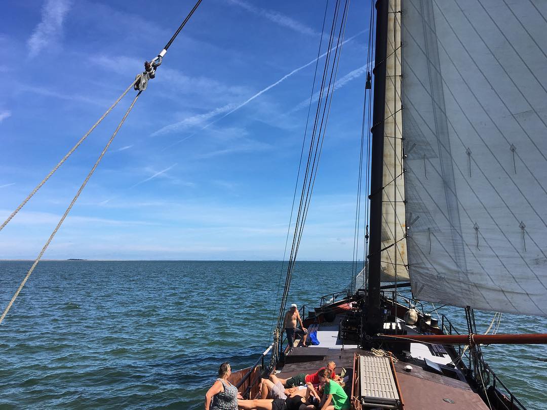 Traditional sailing ship on open water with people relaxing on deck under a bright blue sky in the Netherlands.
