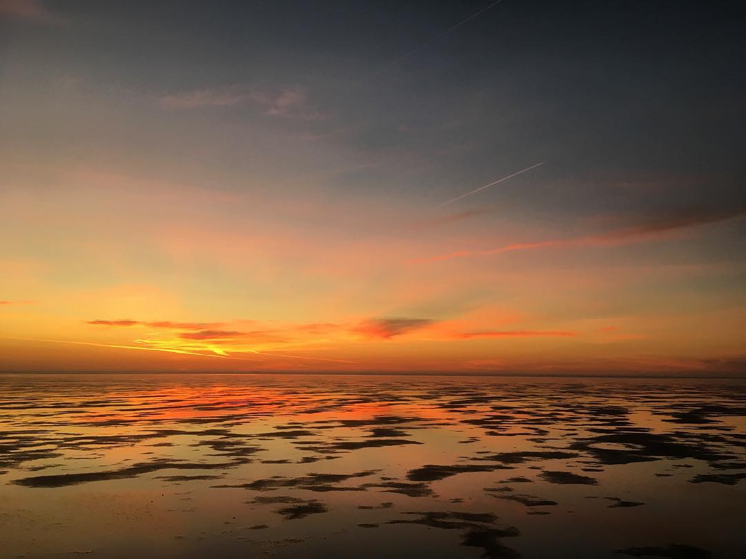 Sunset over wet, reflective tidal flats in the Dutch Wadden Sea, glowing orange in the evening light.