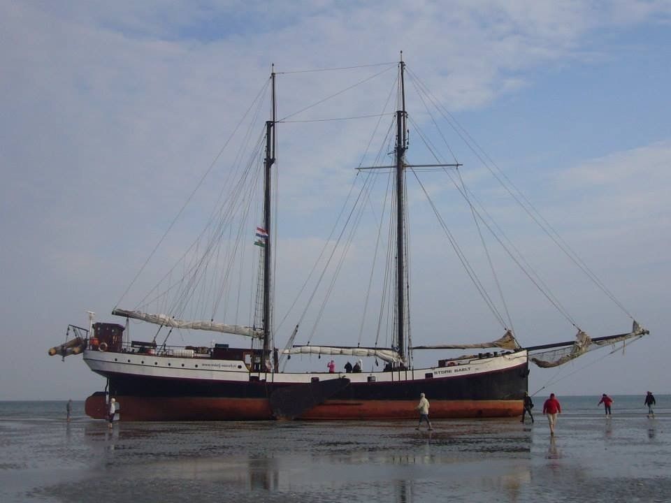 Large traditional sailing ship dried out on the Wadden Sea mudflats, with people walking past at low tide.