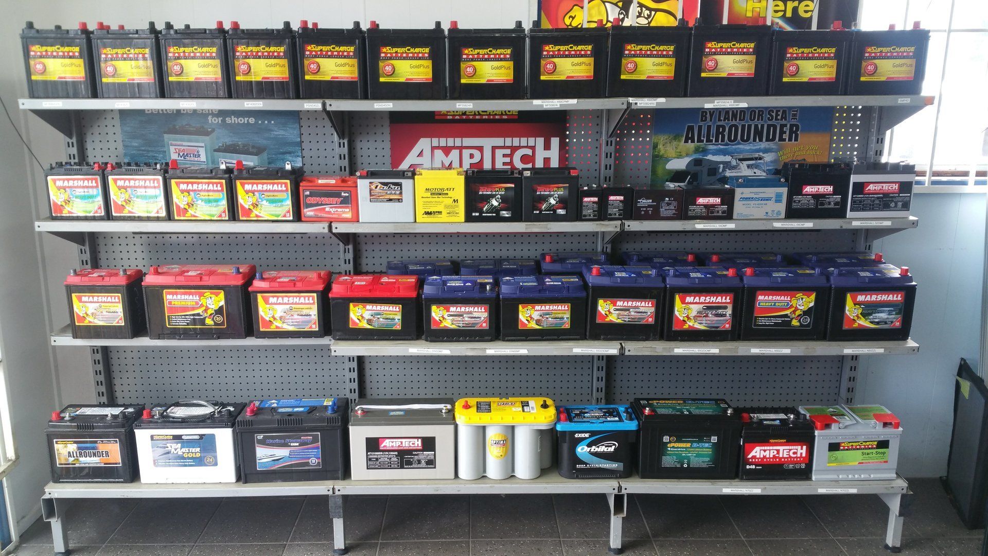 Rows of car batteries on store shelves, with AMIPEC and AMARON signage in the background.