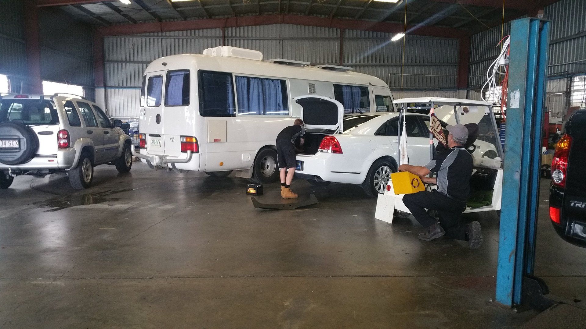 Vehicles parked inside a wet garage, with people working on cars under a metal roof.