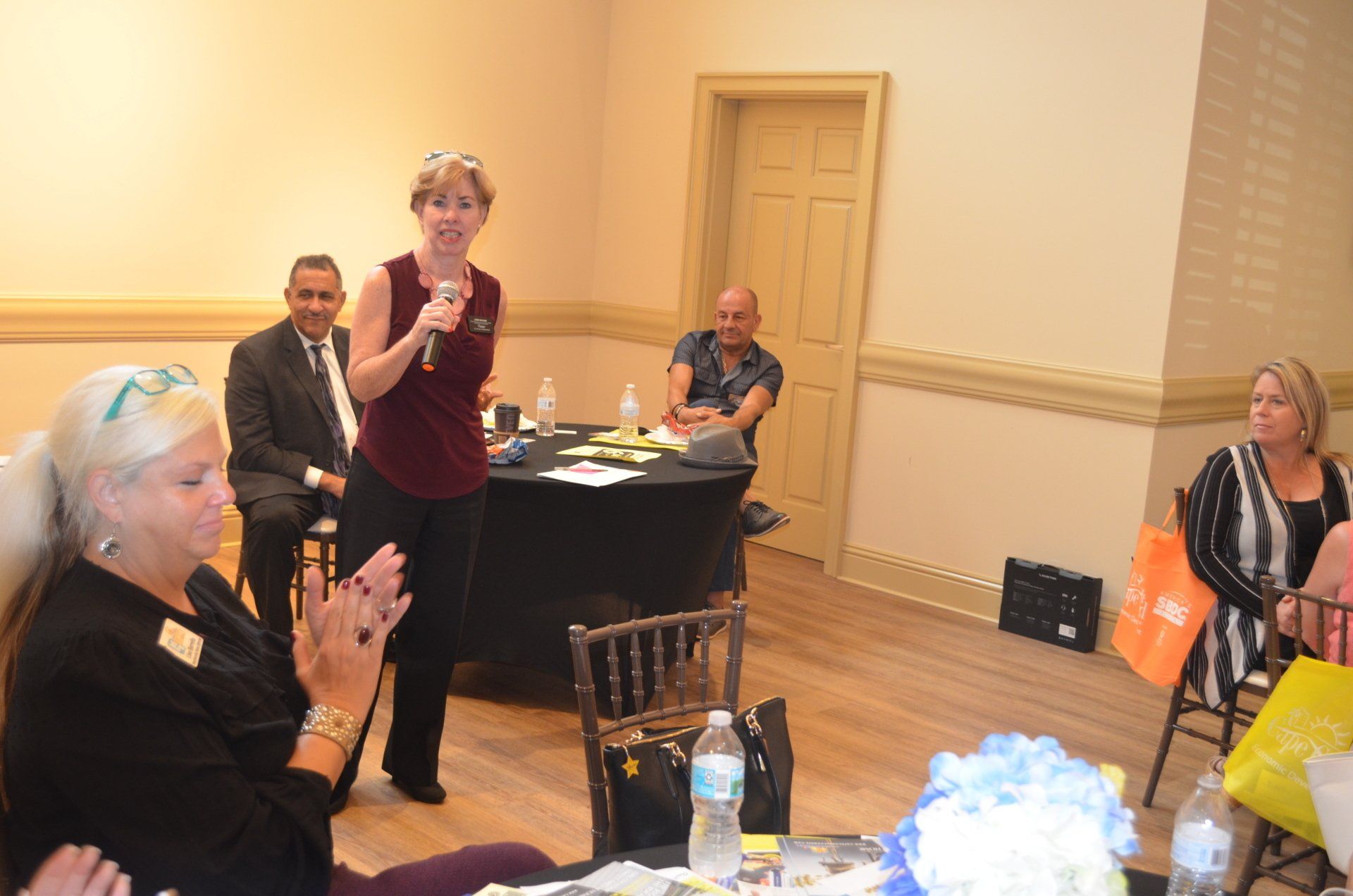 Woman speaking at an event, holding a microphone, with seated audience in a room with a table.