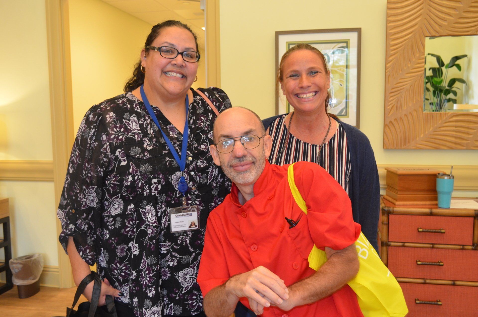 Three people smiling, posed together indoors. One seated in a red shirt, flanked by two others.