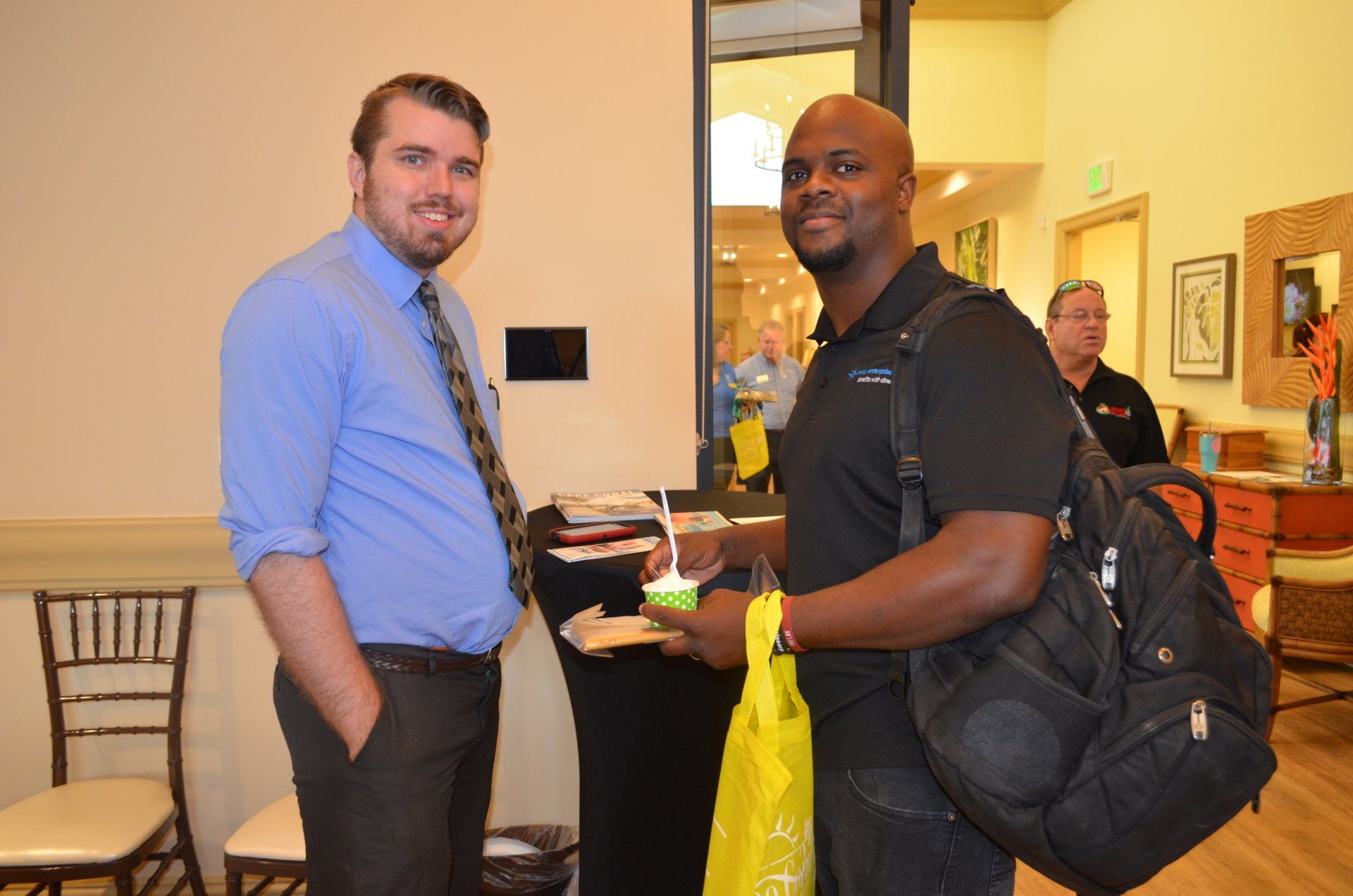 Two men standing indoors, smiling. One in a blue shirt and tie, the other in a black shirt, holding a bag.