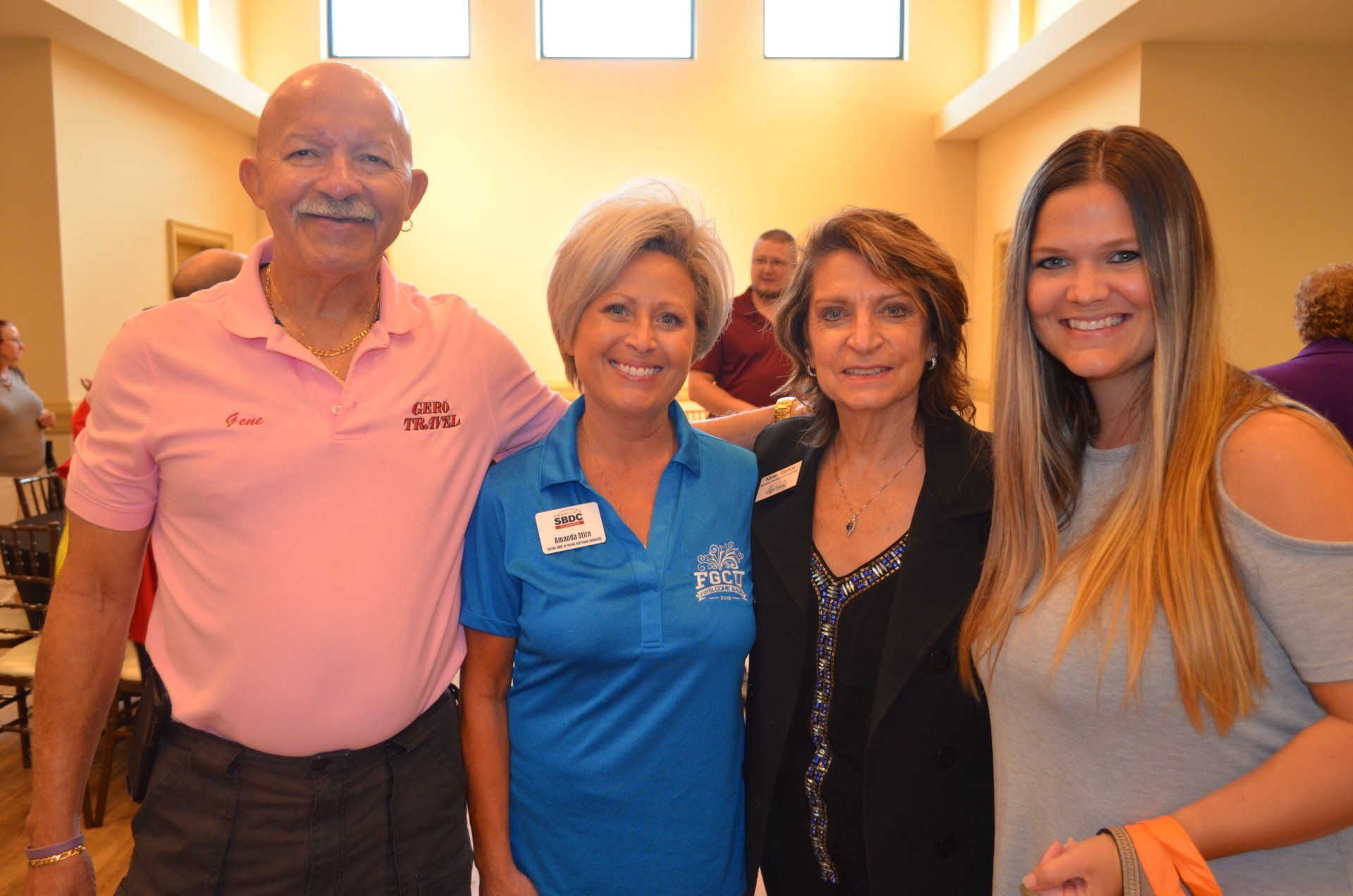 Four people smiling, posing together indoors. A man in pink shirt, a woman in blue, another woman in a black blazer, and a woman with long hair.