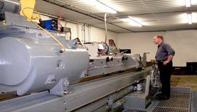 Man observing a large industrial machine in a workshop. The machine is gray, and the setting is brightly lit.
