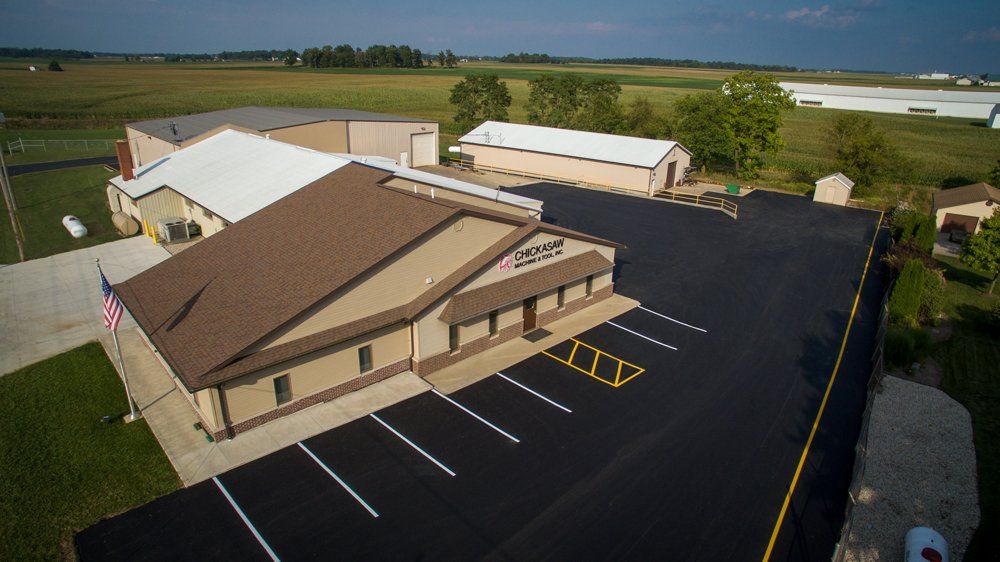 Aerial view of a business with a brown roof and a large, black parking lot in a rural setting.