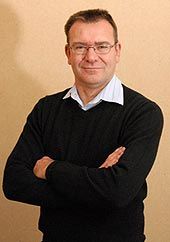 A person wearing glasses, a light-colored collared shirt, and a dark sweater, standing with arms crossed against a tan wall.