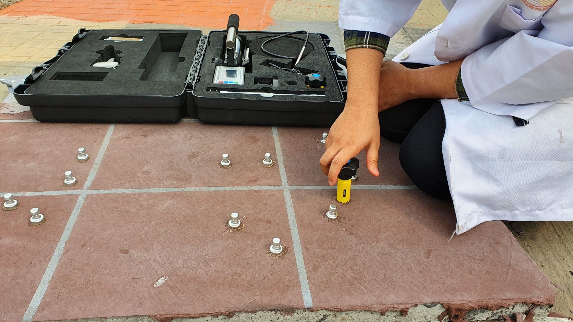 Person in lab coat using a measuring tool on a tiled surface with small silver objects. A black case sits nearby.