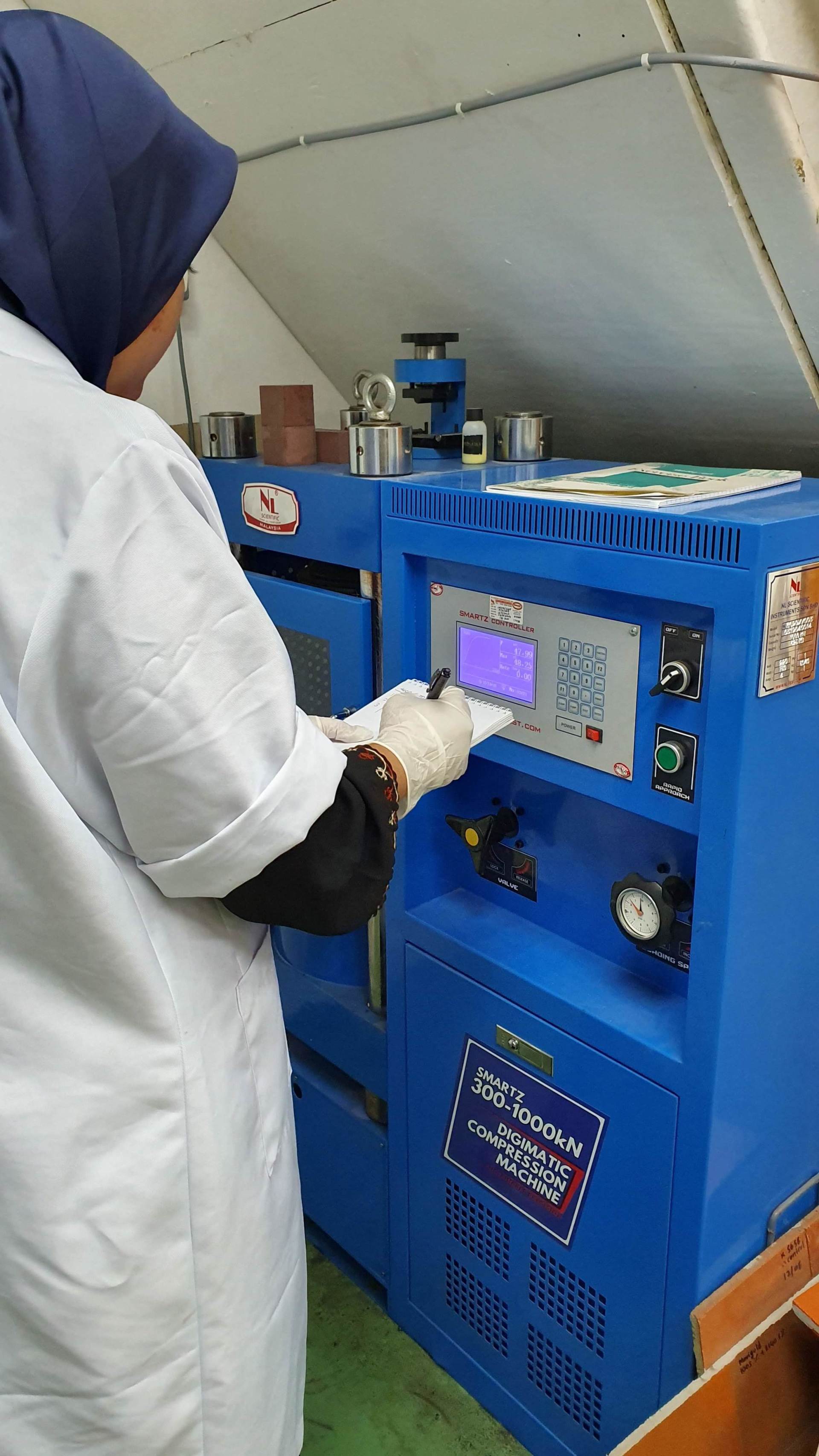 Woman in lab coat operating a blue industrial machine, wearing a hijab and gloves.