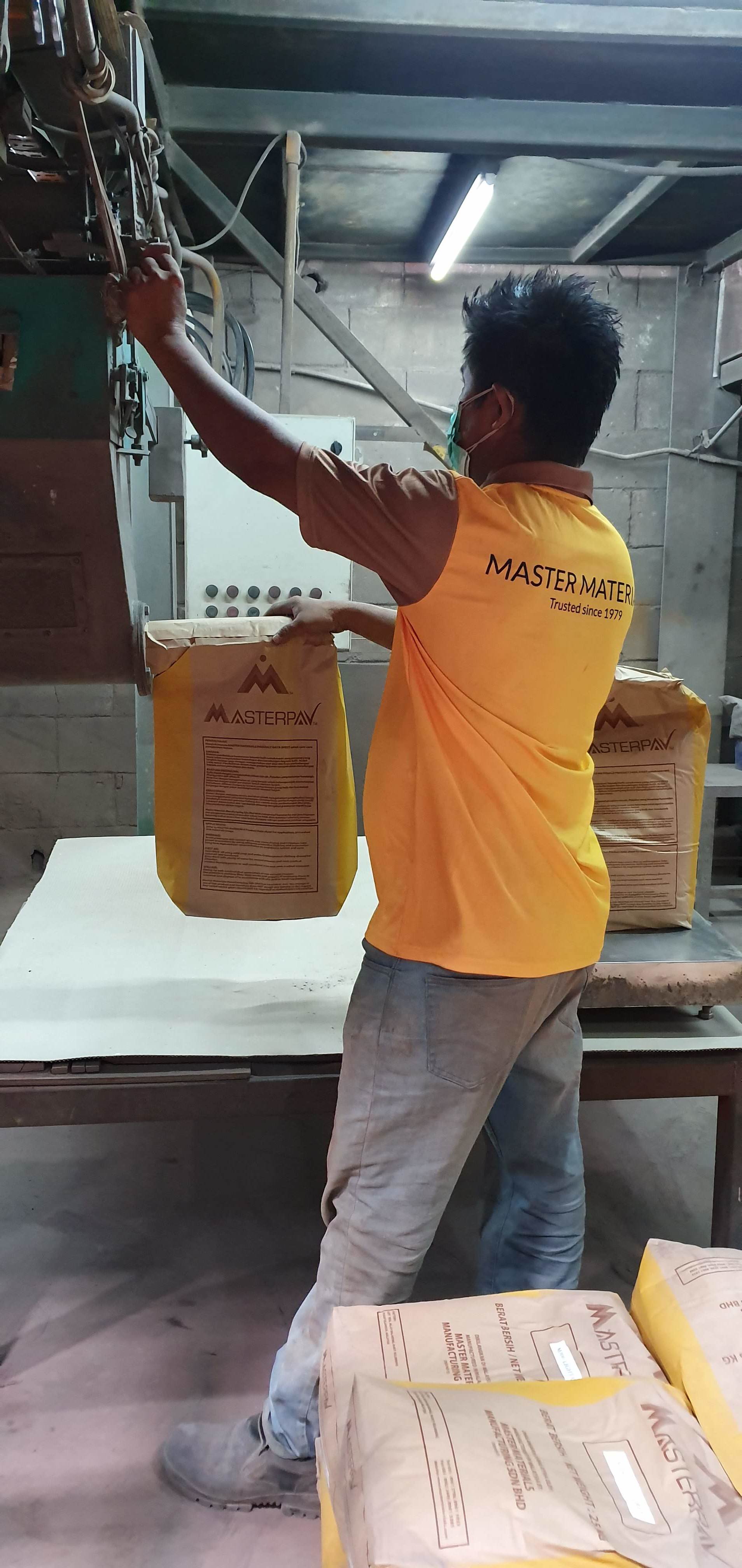 A person in a yellow shirt at a factory holding a bag of product near a machine.