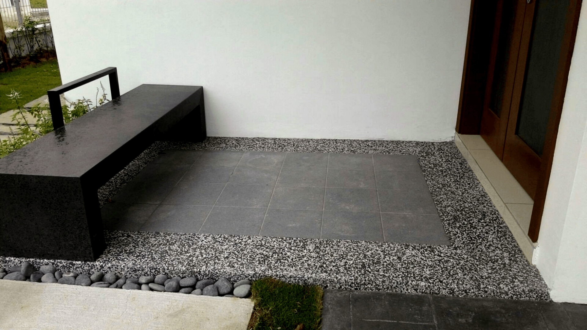 Outdoor seating area with gray bench, stone tiles, and pebbles against a white wall and wooden door.