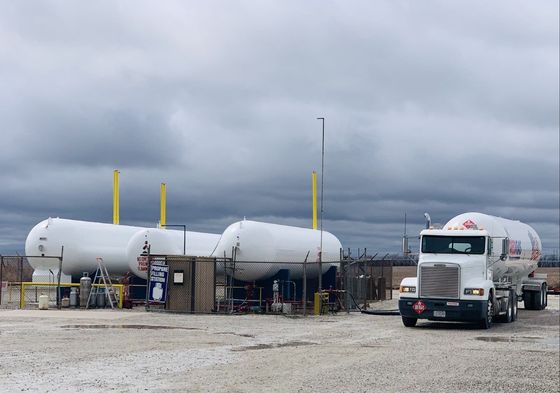 A white tanker truck at a propane storage facility with large white tanks under a cloudy sky.