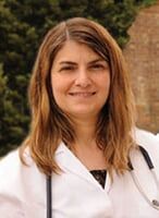 Woman with long brown hair, wearing a white coat and stethoscope, smiling outdoors.