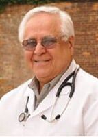 Smiling doctor with glasses, stethoscope, and white coat against a brick wall.