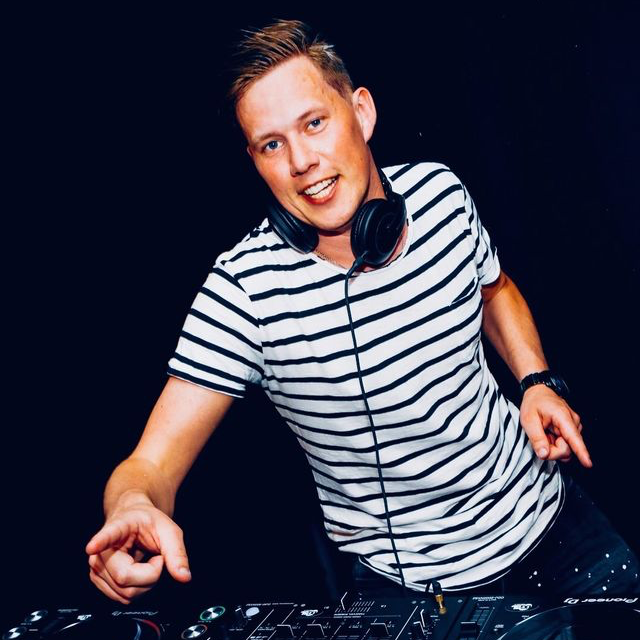 Een dj in een gestreept shirt met koptelefoon op staat voorovergebogen achter een draaitafel, tegen een donkere achtergrond.