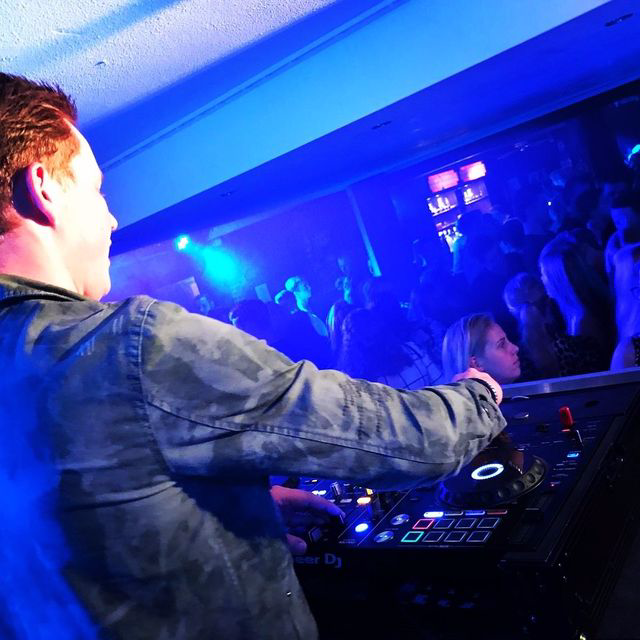 DJ draait in een clubbooth onder blauw licht, met uitzicht op een volle dansvloer.