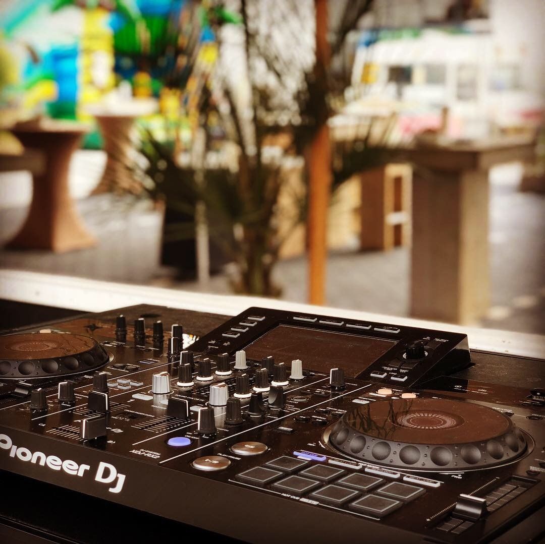 Zwarte Pioneer DJ-controller op een houten tafel in een lichte, sfeervolle buitenruimte van een café.