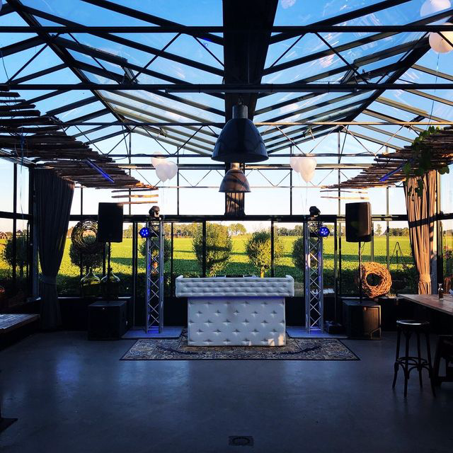 Leeg podium met dj-booth, luidsprekers en lichtslingers onder een glazen paviljoen.