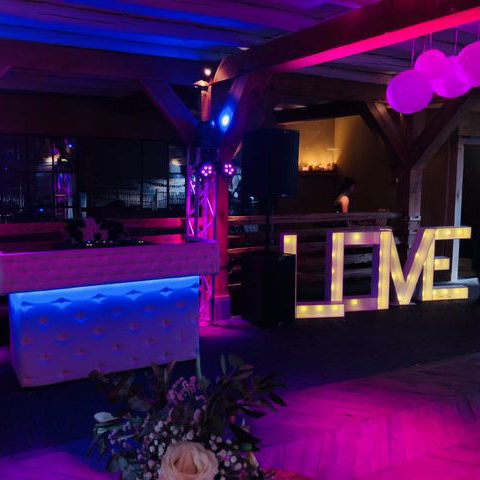 Een schemerig verlichte evenementenlocatie met paarse verlichting, bloemversiering en een oplichtend uithangbord met de tekst 