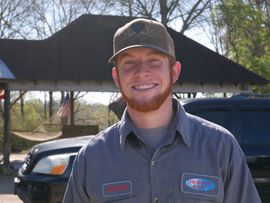 A smiling redhead wearing a work shirt and cap poses outdoors in front of a shaded area and a parked car.